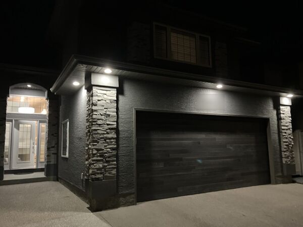 Dark grey stucco house with white stacked stone columns and a wood-look garage door, lit at night